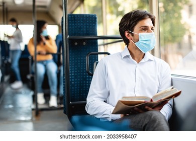 Portrait of pensive man in disposable face mask holding book, going home or to work by bus, looking aside through window and thinking. Pensive guy reading fiction or educational literature
