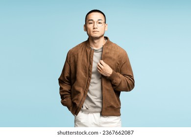 Portrait of pensive serious Asian man wearing stylish brown jacket and eyeglasses, hands in pockets posing isolated on blue background. Advertisement concept - Powered by Shutterstock