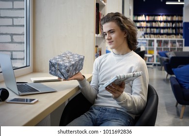 Portrait Of Pensive Student Sitting At Table In Front Of Generic Portable Computer, Holding Copybook And Pen, Writing Composition For Essay Contest, Having Deep In Thoughts Expression On His Face