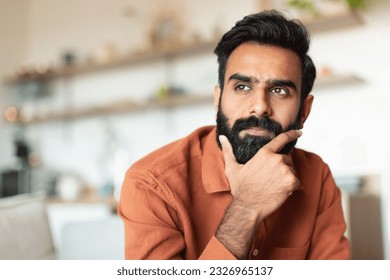 Portrait Of Pensive Young Arabic Man Thinking Sitting In Living Room At Home, Looking Aside. Thoughtful Middle Eastern Guy Touching Beard And Pondering About Issues, Free Space