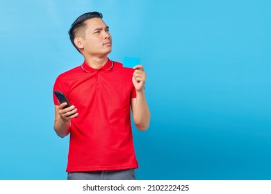 Portrait Of Pensive Young Asian Man Holding Mobile Phone And Credit Card Isolated On Blue Background