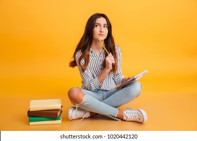 Portrait of a pensive young girl making notes while sitting with books isolated over yellow background - Powered by Shutterstock