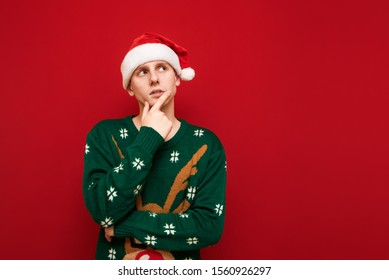 Portrait of pensive young man in santa hat and christmas sweater stands on red background and looks away at blank space and thinks. Thoughtful guy in Christmas is isolated. Copy space - Shutterstock ID 1560926297
