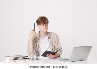 Portrait Of Pensive Young Man Student Wears Beige Shirt And Glasses Thinking And Blowing Bubbles With Chewing Gum At The Table With Laptop Computer And Notebooks Isolated Over White Background