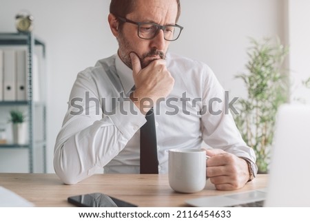 Portrait of perturbed businessman with cup of coffee in the office looking at laptop computer screen and thinking, selective focus