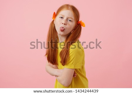 Portrait of petite freckles red-haired girl with two tails, looks and shows tounge at the camera, wears in yellow t-shirt, stands over pink background.