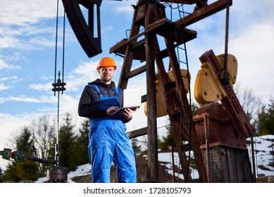 Portrait of petrolium engineer in blue overalls and orange helmet, with laptop, standing with his back to an oil rig, checking oil pumping unit, making notes in his computer, smiling to the camera.
