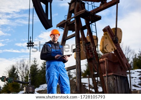 Portrait of petrolium engineer in blue overalls and orange helmet, with laptop, standing with his back to an oil rig, checking oil pumping unit, making notes in his computer, smiling to the camera.