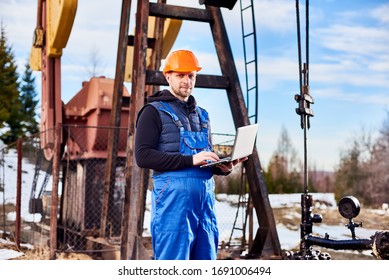 Portrait of petrolium engineer, wearing blue overalls and orange helmet, with a laptop, standing with his back to an oil rig, checking oil pumping unit, making notes in his computer