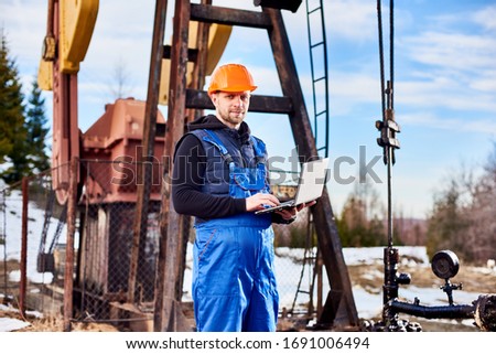 Portrait of petrolium engineer, wearing blue overalls and orange helmet, with a laptop, standing with his back to an oil rig, checking oil pumping unit, making notes in his computer