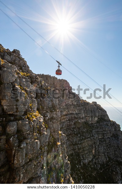 Portrait photo of the cable car going up a rocky hill\
with steep cliffs, Lion\'s Head, the ocean and a city in the\
background. Shot in Table Mountain, Cape Town, Western Cape, South\
Africa. 