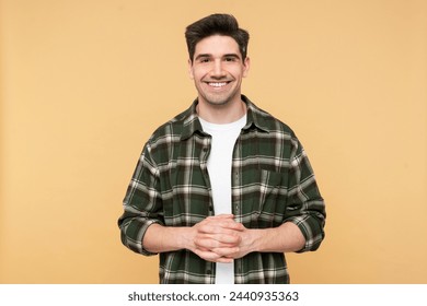 Portrait photo of a cheerful, bearded man smiling joyfully with intertwined fingers, wearing a plaid shirt, posed against an isolated yellow background, looking at the camera - Powered by Shutterstock