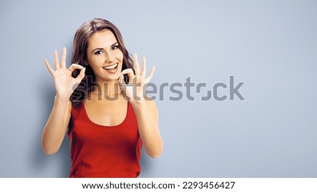 Portrait photo - happy smiling beautiful woman in casual clothing, showing okay ok gesture, or zero hand sign, over grey color wall background. Girl in red dress. Brunette excited model at studio.