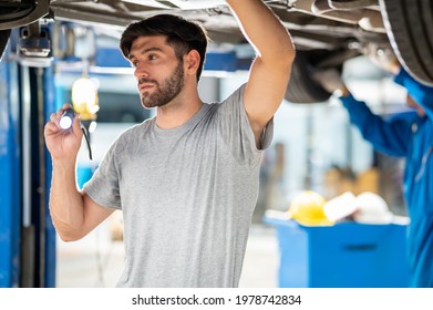 Portrait Photo Of Professional Look Caucasian Vehicle Service Technician Standing Under Car Checking Vehicle Under Body And Holding Lighting Torch In Car Repair Shop.