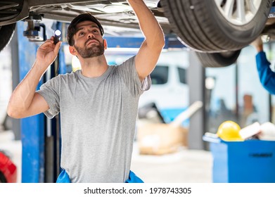 Portrait Photo Of Professional Look Caucasian Vehicle Service Technician Standing Under Car Checking Vehicle Under Body And Holding Lighting Torch In Car Repair Shop.