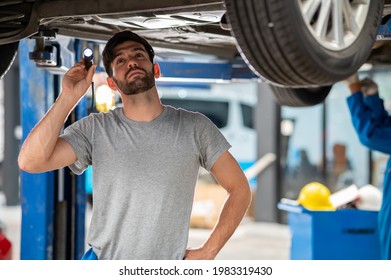 Portrait Photo Of Professional Look Caucasian Vehicle Service Technician Standing Under Car Checking Vehicle Under Body And Holding Lighting Torch In Car Repair Shop.