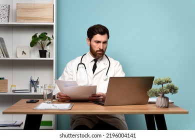 Portrait Photo Of Serious Family Doc Man Wearing White Lab Coat And Stethoscope Sitting And Looking Laptop Screen While Working At Cabinet With Blue Walls 