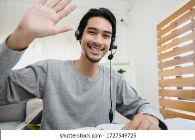 Portrait photo of smart Asian guy using video call communicate with business colleague friend from home during self isolation from coronavirus outbreak crisis. Asian man waving hand looking at camera.