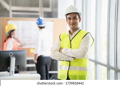 Portrait Photo Of Young Smart Hispanic  Engineer Wearing Safety Helmet Standing Indoor Of Company Office Or Construction Site With Colleague Discussing About Work In Distance.