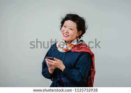 Portrait of a photogenic middle-aged Chinese Asian woman in a blue dress and colorful scarf texting on her smartphone. She is smiling and is happy as she texts and has short hair. 