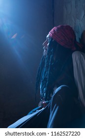 Portrait The Pirate With Black Dreadlocks, A Red Bandana And In A White Shirt With A Long Sleeve, A Blue Moonlight. Cinema Atmosphere. Dark Background