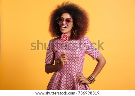 Portrait of a playful afro american woman in retro style clothes holding lollipop and looking at camera isolated over yellow background