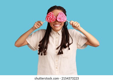 Portrait of playful childish romantic woman with black dreadlocks covering eyes with sugary hear shape candies, wearing white shirt. Indoor studio shot isolated on blue background.