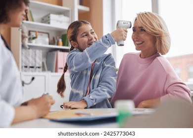 Portrait of playful girl having fun at doctors office and measuring moms temperature with contactless thermometer - Powered by Shutterstock