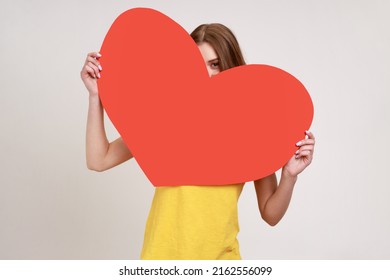 Portrait Of Playful Teenager Girl Peeking Out Of Big Read Heart With Flirting Look And Expressing Love, Greeting On Valentine's Day. Indoor Studio Shot Isolated On Gray Background.