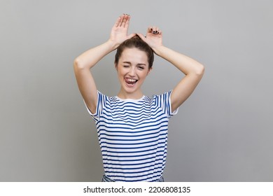 Portrait Of Playful Woman Wearing Striped T-shirt Making Funny Bunny Ears With Hands On Head, Childish Behavior, Optimistic Mood. Indoor Studio Shot Isolated On Gray Background.