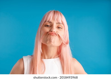 Portrait of playful young woman with natural long pink dyed hair holding a strand of hair as a moustache, posing isolated over blue studio background. Beauty, hair care concept