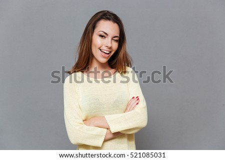Portrait of a playful young woman in sweater standing with arms folded and winking isolated on the gray background