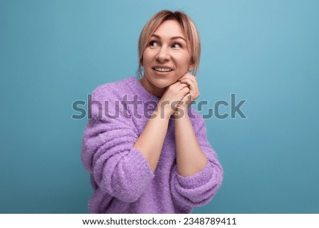 portrait of a pleasant charming pleasant blond young woman in a purple sweater smiling on a blue background with copy space