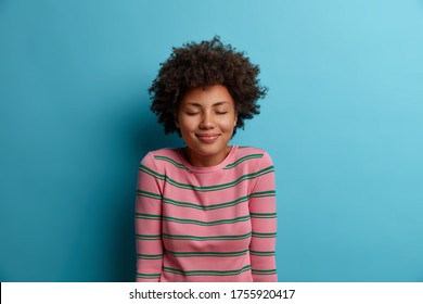 Portrait of pleasant looking dark skinned woman stands with eyes shut, being in good mood, imagines something nice, dreams about perfect vacation, wears striped sweater, isolated on blue background