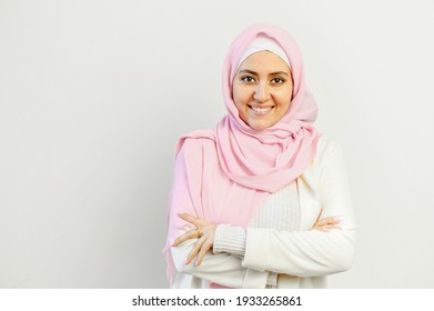 Portrait of a pleasant Muslim Arabic mixed-race businesswoman entrepreneur or successful business owner in pink hijab standing with the arms folded. smiling and posing against gray wall background