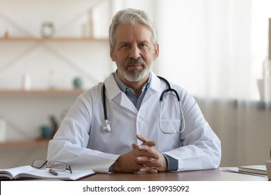 Portrait Of Pleasant Professional Older Mature Gp Physician In White Coat Sitting At Table, Waiting For Patient. Smiling Trusted Middle Aged Male Doctor Looking At Camera, Medical Insurance Service.