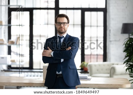 Portrait of pleasant young 30s male ceo executive manager in formal wear standing with folded arms in modern office, showing confidence and leadership qualities, feeling proud of corporate success.