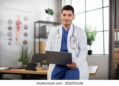 Portrait of pleasant young Arabian doctor in white coat and blue scrubs, with stethoscope around neck, leaning on the table in modern office and using laptop pc for medical records or prescribing