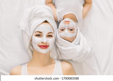 Portrait of pleasant young mother and her little daughter lying together on bed with white towels on head and facial clay mask while enjoying spa procedures. Isolated, top view. Beauty concept - Powered by Shutterstock