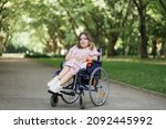 Portrait of pleasant young woman with spinal muscular atrophy smiling on camera among green summer park. Female person who using wheelchair. Concept of people with disability.