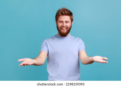 Portrait Of Pleased Bearded Man Sharing Opening Hands Looking At Camera With Kind Smile, Greeting And Regaling, Happy Glad To See You. Indoor Studio Shot Isolated On Blue Background.