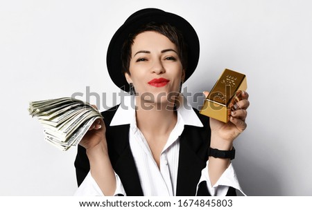 Portrait of pleased lucky woman in stylish hat, black jacket and white shirt holding a fan of dollars and a bar of gold in her hands sending us a kiss over white background
