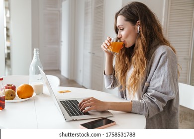 Portrait of pleased woman drinking juice and using earphone while working with laptop in bright kitchen - Powered by Shutterstock