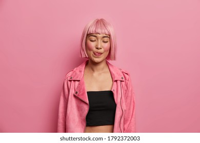 Portrait of pleased young Asian woman licks lips, stands with closed eyes, remembers pleasant taste of something, wears black top and pink jacket, poses against rosy background, feels satisfied