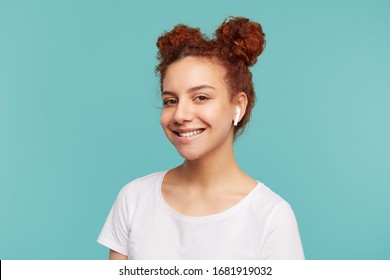 Portrait of pleased young brown-eyed curly brunette woman with earphones looking positively at camera with charming smile while standing over blue background