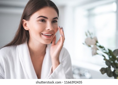 Portrait Of Pleased Young Lady Touching Cheek With Hand. She Putting Cream On Face Skin Indoor