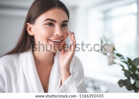 Portrait of pleased young lady touching cheek with hand. She putting cream on face skin indoor