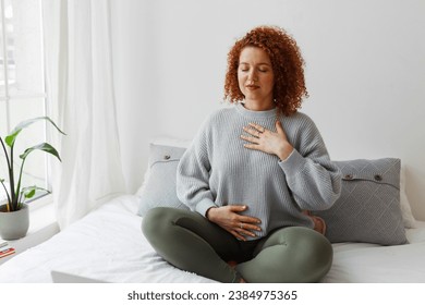 Portrait of plus-size curly redhead female in casual clothes doing pranayama technique sitting on bed next to window with closed eyes, putting hands on chest, breathing slowly and deeply - Powered by Shutterstock