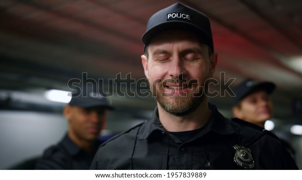 Portrait of police officer smiling at camera. Male\
cop wearing cap and police uniform. Positive policeman posing at\
camera. Colleagues standing on background with flashing lights of\
patrol car