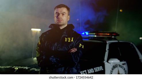 Portrait of police officers with serious faces looking at camera. FBI agent work at the scene at night, police car with lights on background.